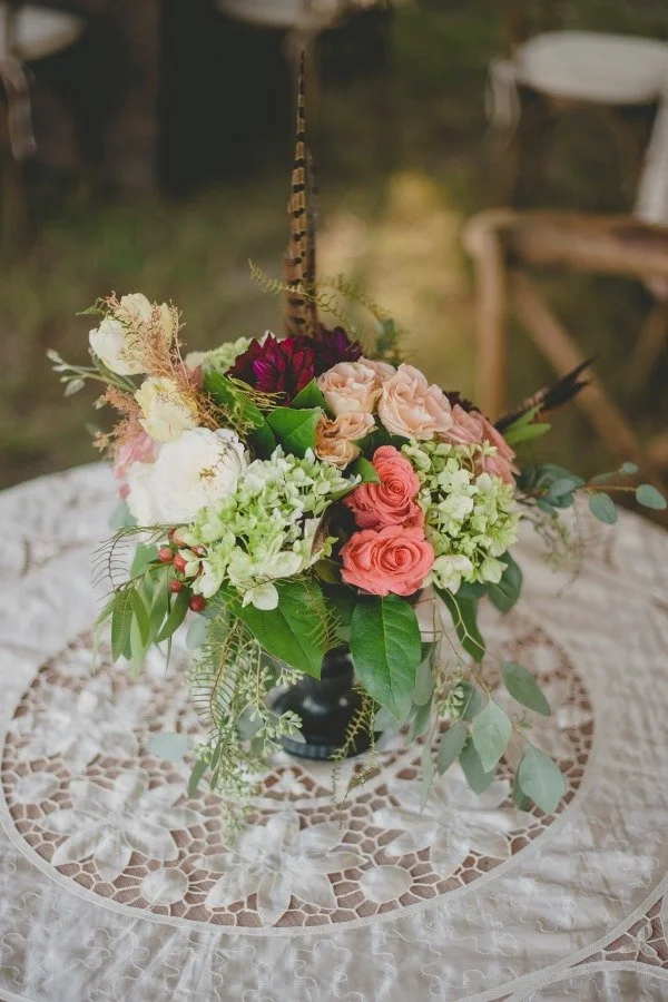 Colorful flower arrangement with pink, white, and dark purple flowers, and green foliage in a black vase, placed on a lace tablecloth.