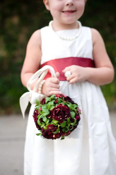 Close-up of a young girl in a white dress with a red sash, holding a small bouquet of dark red and green flowers, outdoors.