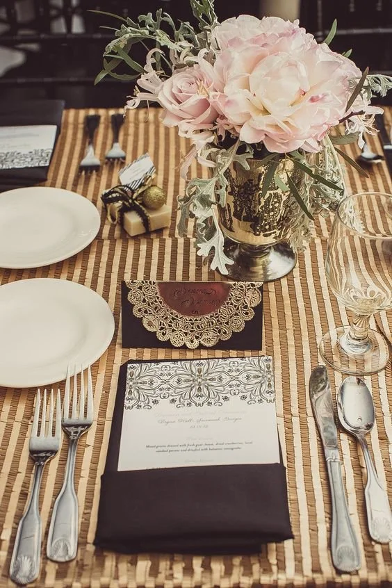Table setting with a floral centerpiece featuring pink roses and greenery, plates, silverware, a glass, a menu, a placed card, and small wrapped gifts on a striped tablecloth.