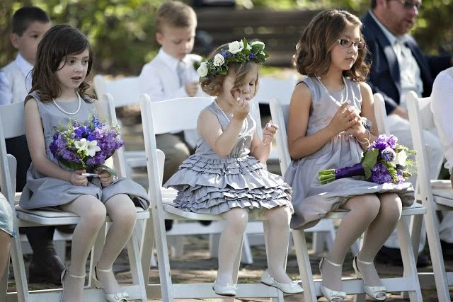 Three young girls in lavender dresses sitting in a row at a wedding ceremony, holding bouquets, with other attendees in the background.