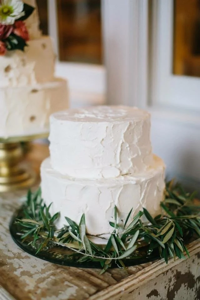 Two-tiered white frosted cake on a black cake board, decorated with green olive branches, sitting on a rustic wooden table.