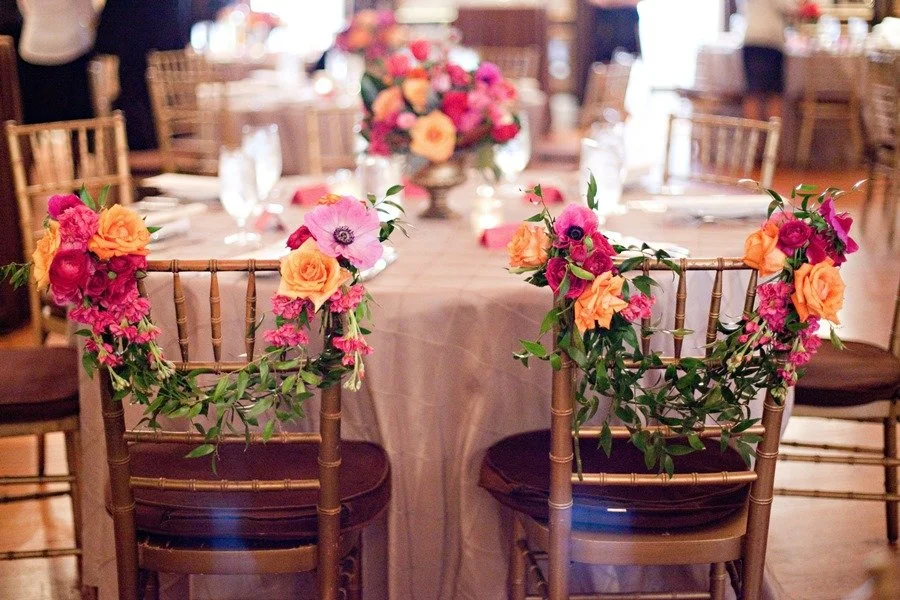 Decorated chairs around a banquet table with floral arrangements, set for an event.