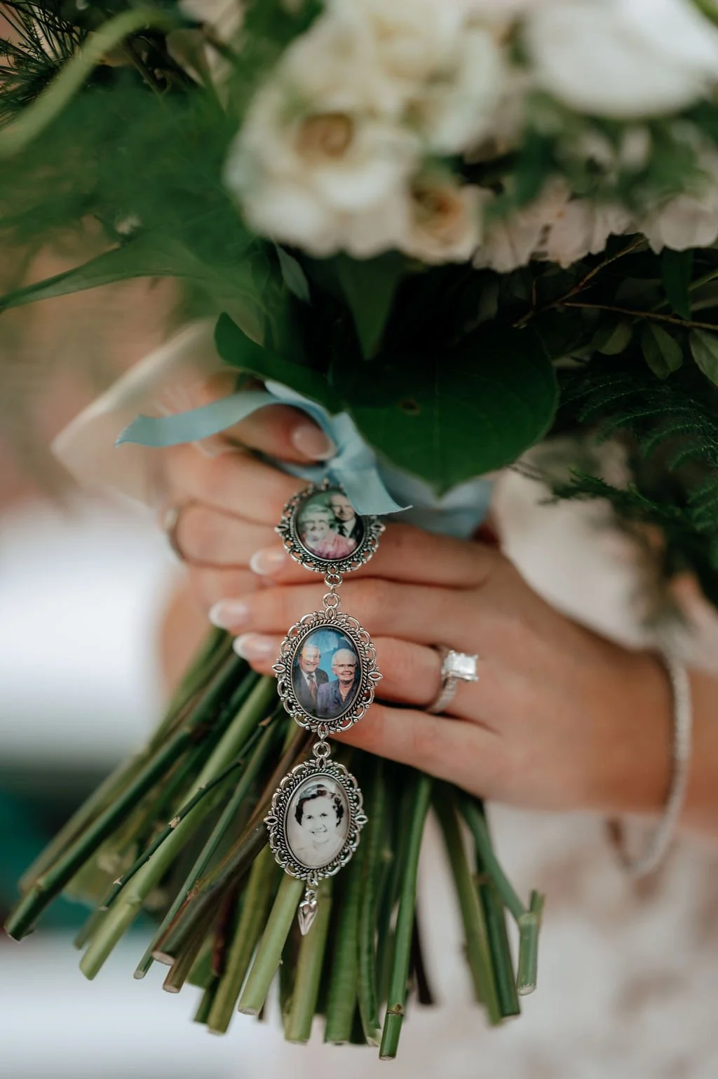 A woman's hand holding a bouquet of white flowers with a blue ribbon. The bouquet has medallion-style photo charms attached, featuring images of elderly people, including a smiling woman. The woman is wearing a ring with a large stone, a bracelet, an