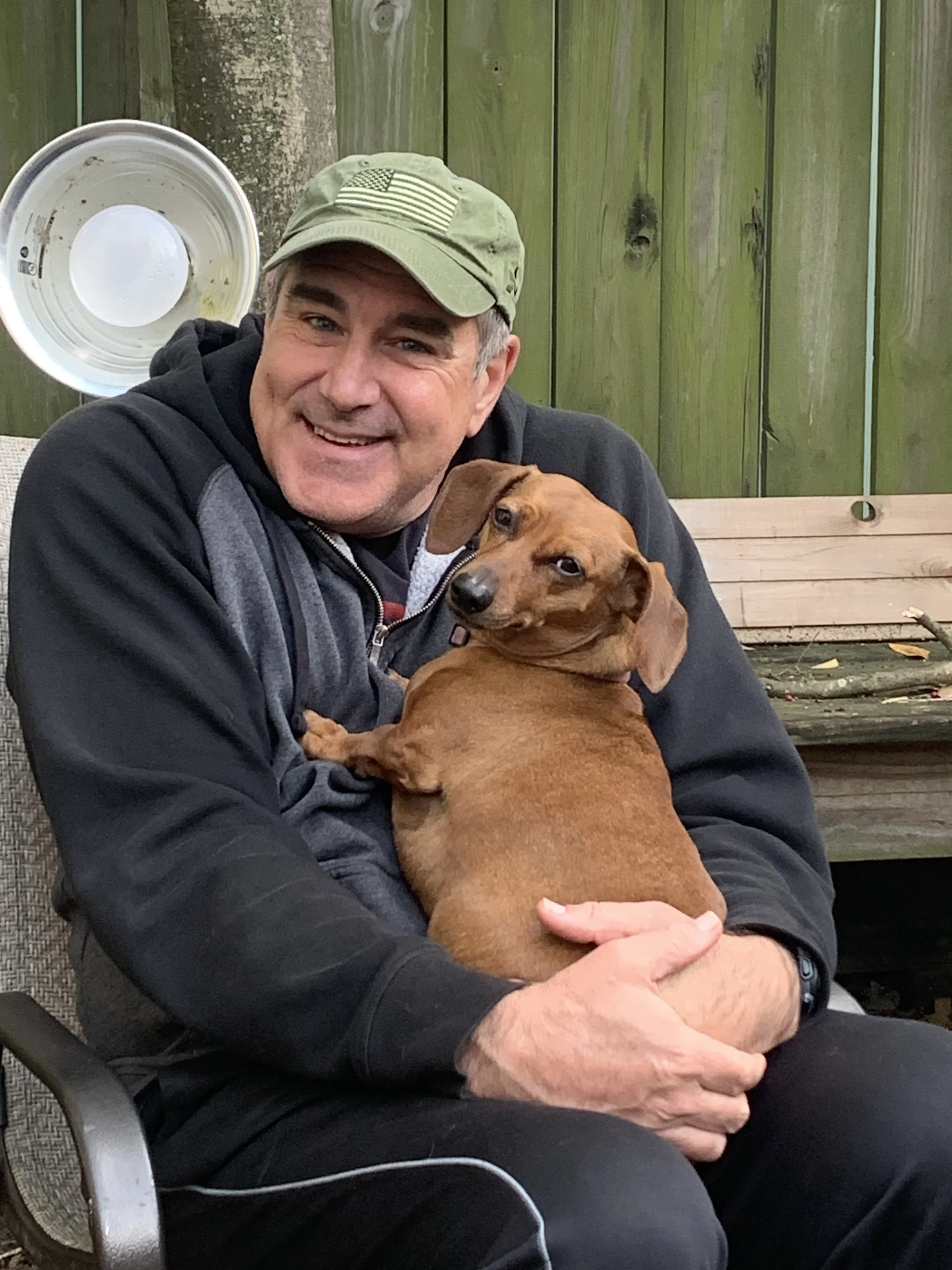 A man wearing a baseball cap with an American flag patch, black hoodie, and black pants, sitting outdoors on a chair, holding a brown dachshund dog. There is a green wooden fence and a tree in the background.