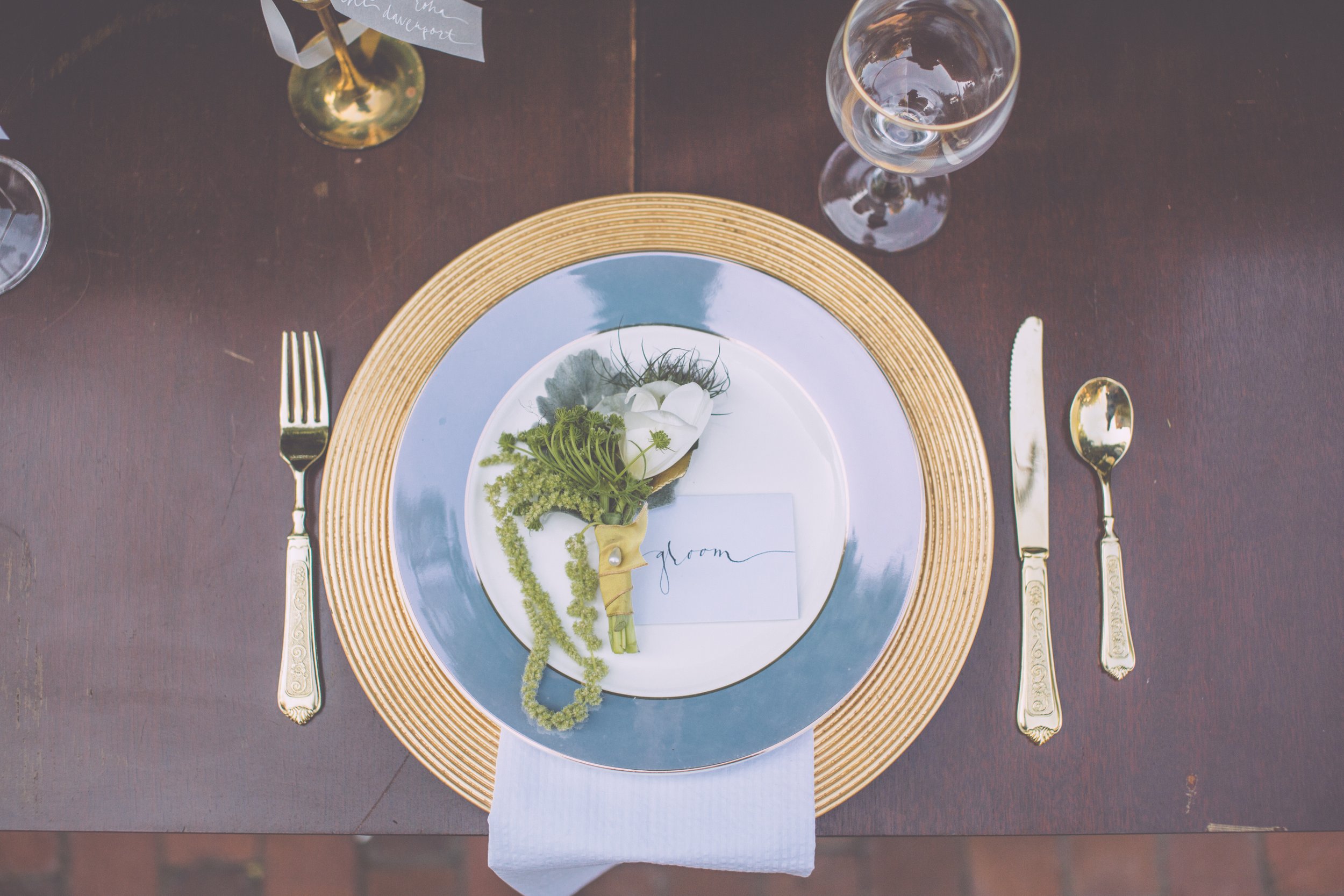 Elegant table setting with gold-rimmed plates, silver flatware, and two wine glasses. Place card labeled 'groom' decorated with flowers and greenery, on a dark wooden table.