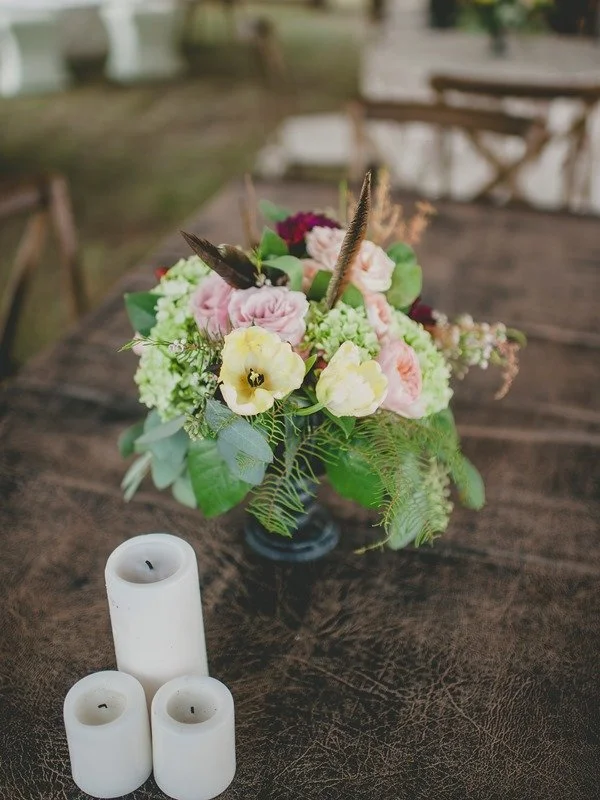 A floral arrangement with pink, yellow, and green flowers and feathers in a dark vase, placed on a wooden table with three white candles nearby.