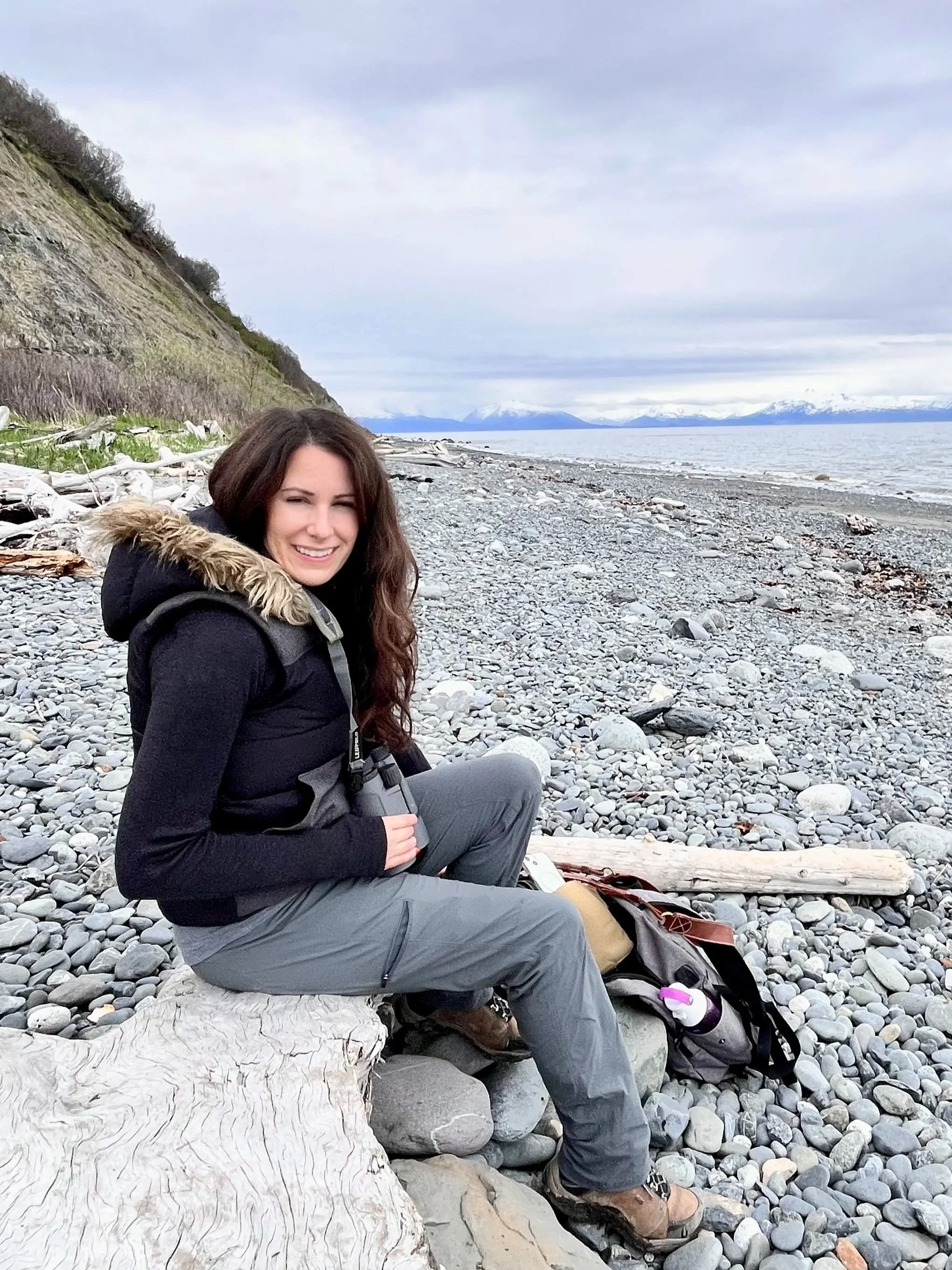 A woman smiling and sitting on a large piece of driftwood on a rocky beach. Behind her, there is a cliffside on the left, water on the right, and snow-capped mountains in the distance under a cloudy sky.