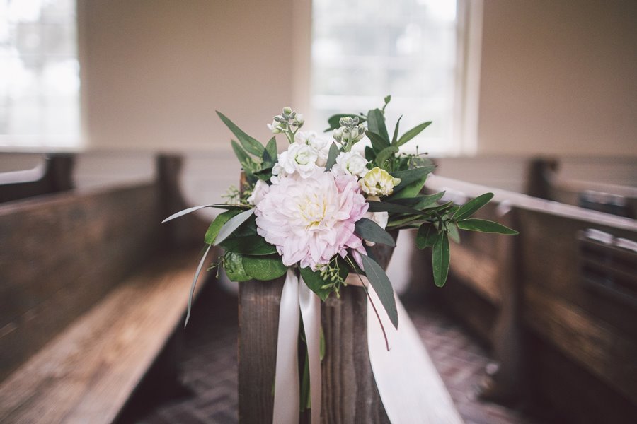 A floral arrangement of white and pink flowers with green foliage in a wooden stand inside a church with wooden pews.