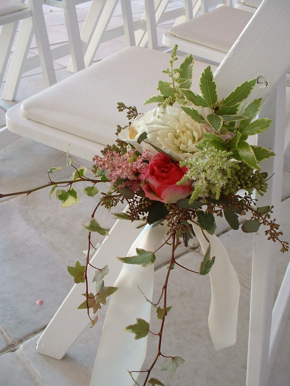 Flower arrangement with white, pink, and green flowers and leaves, tied to a white chair with a ribbon, on a patio with white chairs.
