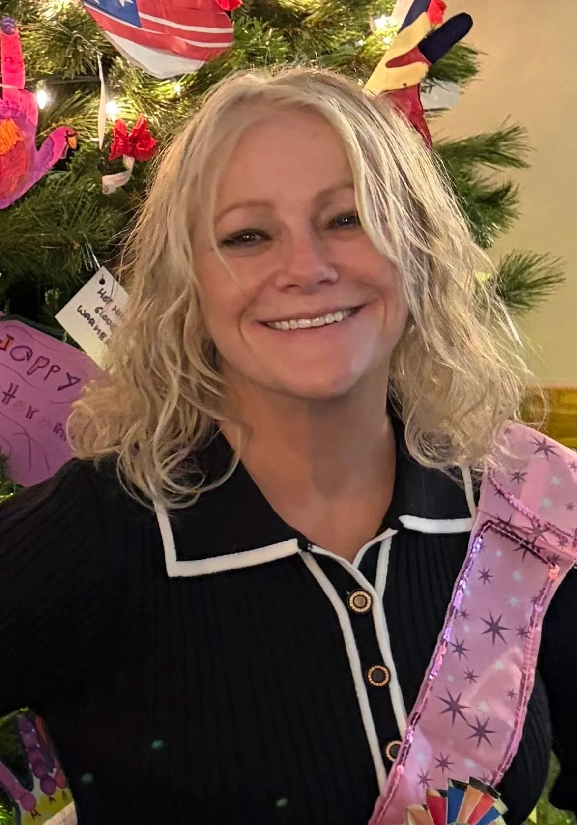 A woman with blonde curly hair smiling in front of a decorated Christmas tree.