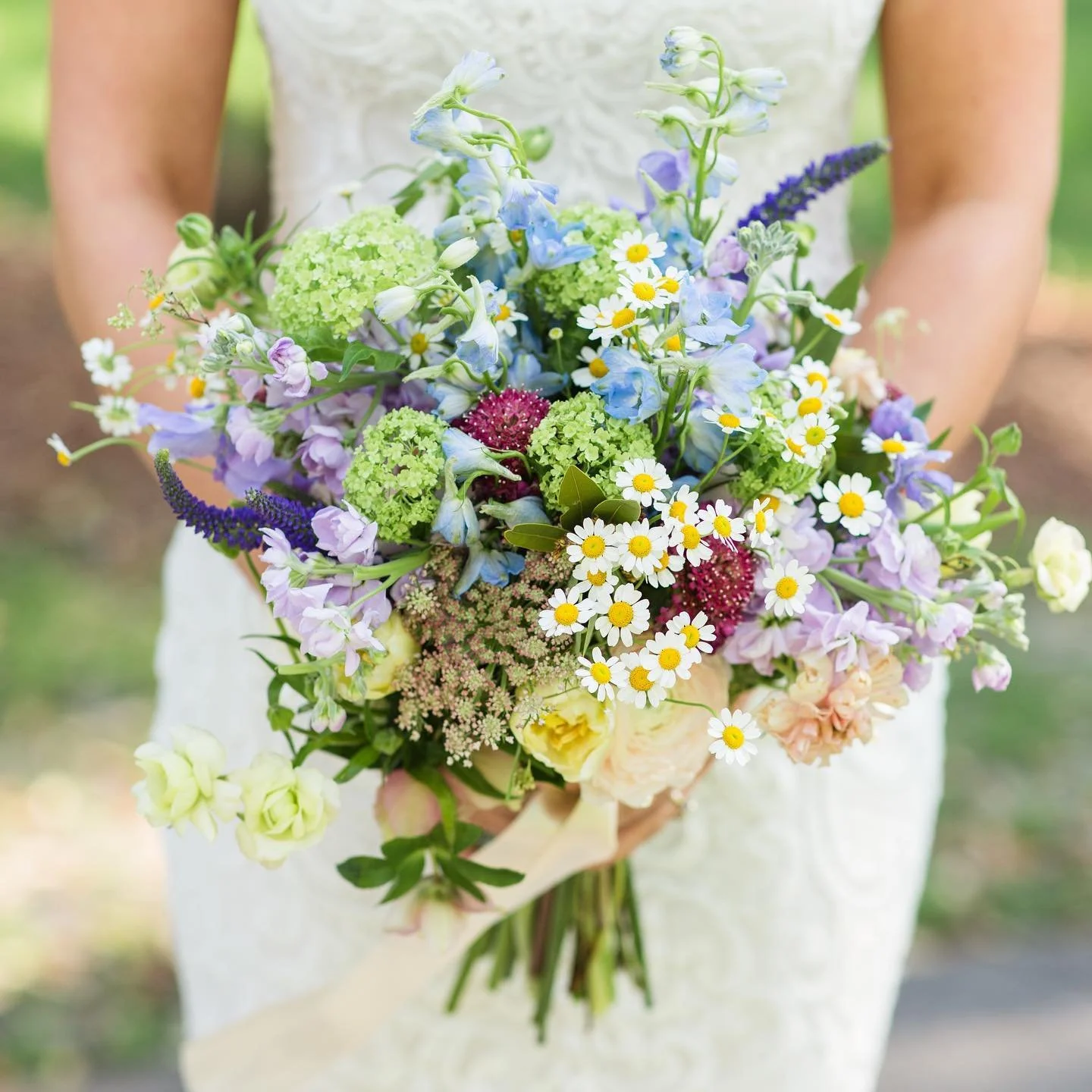 A person holding a colorful bouquet of mixed flowers, including daisies, blue delphiniums, purple and green hydrangeas, and other assorted blooms, against a blurred outdoor background.