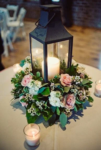 A candle lantern surrounded by a floral arrangement with pink and white flowers and green leaves, placed on a table with smaller candles.