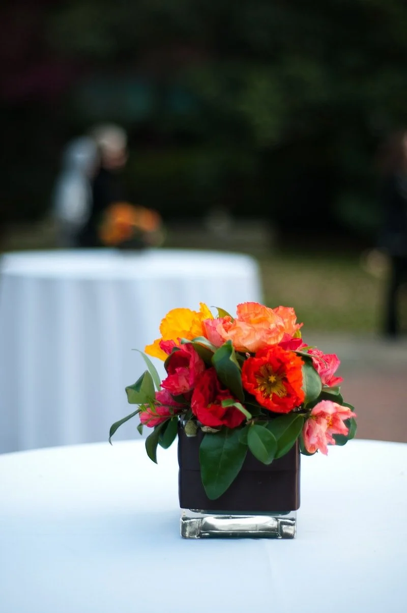 A colorful flower arrangement in a black square vase on a white table, with blurred outdoor background and other tables.