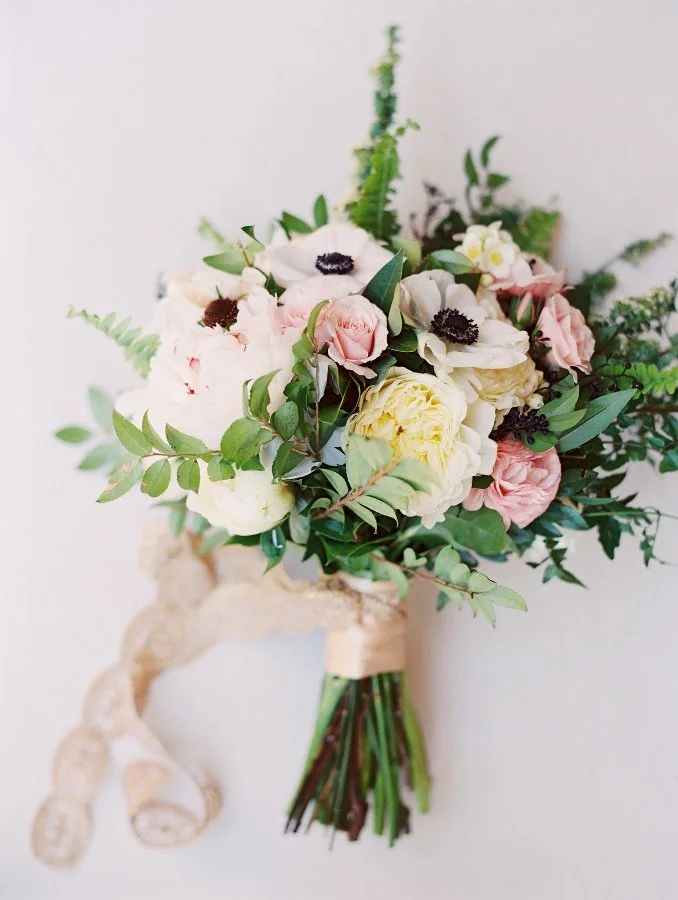 A bouquet of white, pink, and yellow flowers with greenery, tied with a beige ribbon, against a light background.