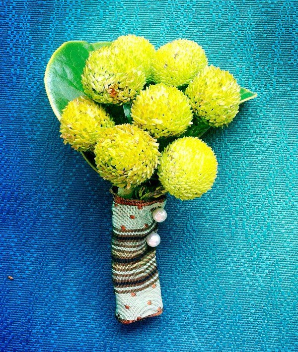 A bouquet of yellow flowers with green leaves, wrapped in fabric with a striped pattern, decorated with small pearl embellishments, on a textured blue surface.