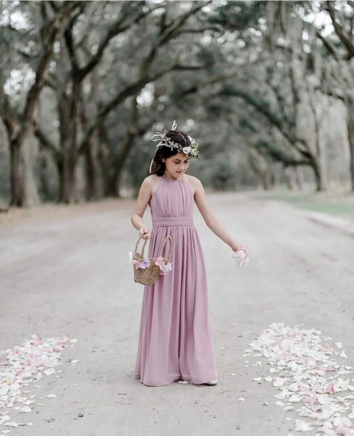 A young girl dressed in a long, pink dress is walking on a dirt path lined with flower petals. She is holding a small basket with flowers and has a floral crown on her head. The background features trees with arching branches and greenery.