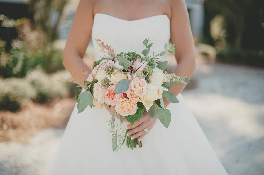 A bride holding a bouquet of pink and cream roses with green leaves outdoors.
