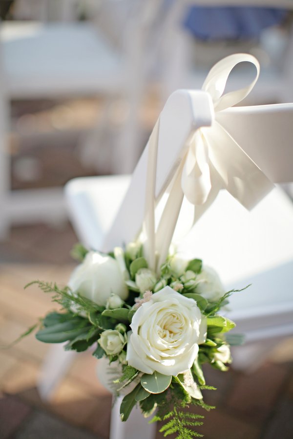 White floral arrangement with roses and greenery attached to a white chair at a wedding or special event.