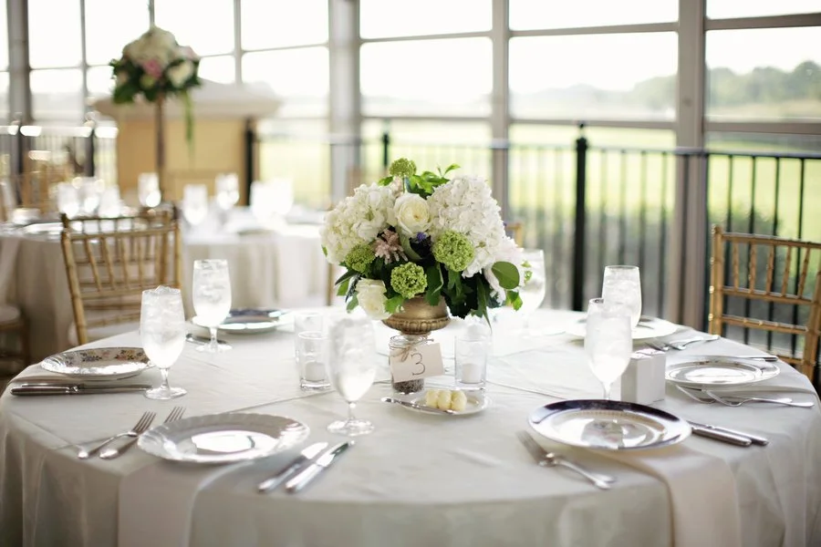 Round banquet table with a white tablecloth, set with plates, silverware, and glasses. Centered is a floral arrangement with white and green flowers in a gold vase, and a small table number '3'. In the background are more tables and chairs with a vie