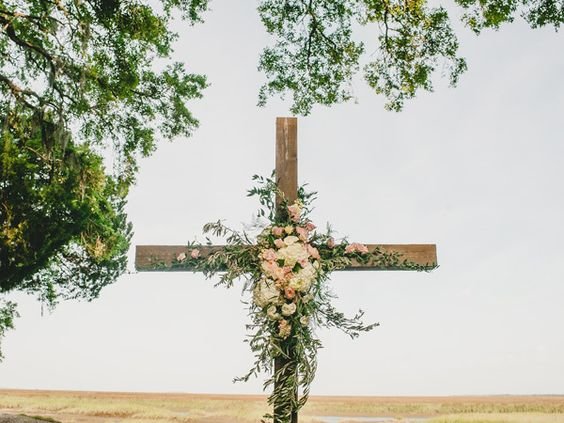 Wooden cross decorated with pink and white flowers, set outdoors with trees in the background.