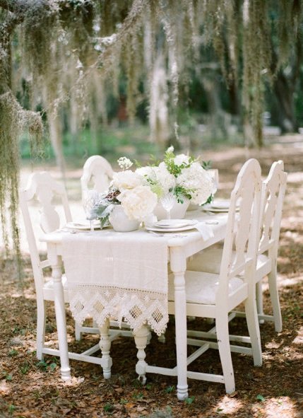 An outdoor dining table set with white flowers and dishes in a wooded area with hanging moss.