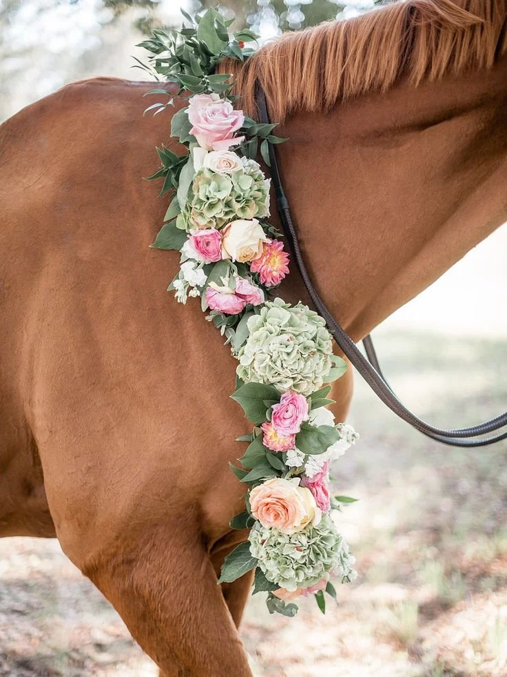 Close-up of a brown horse wearing a floral garland with pink, white, and peach roses, hydrangeas, and greenery around its neck.