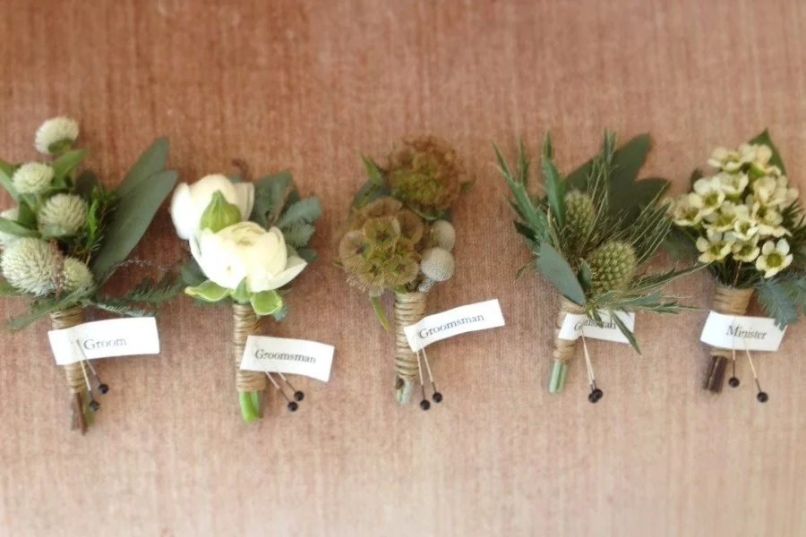 Five small boutonnieres with different flower arrangements, labeled from left to right: Groomsman, Groomsman, Groomsman, Groomsman, Minister, all placed on a wooden surface.