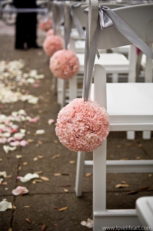 Pink pom-pom decorations hanging from a white chair at an outdoor wedding or event venue, with flower petals scattered on the ground.