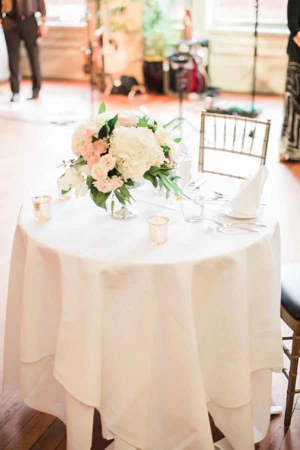 Round table with white tablecloth, set with silverware, glasses, white napkins, and a floral centerpiece of white and pink flowers with green leaves, in a well-lit indoor venue.