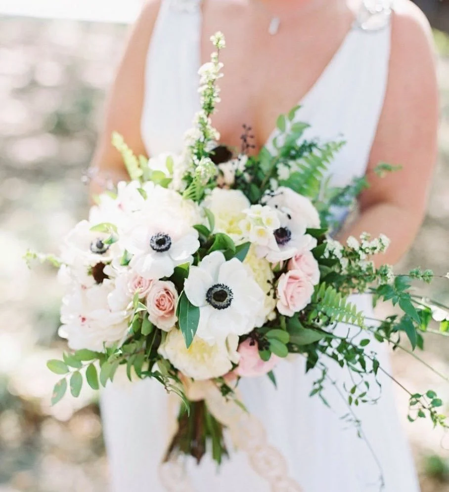 A bride holding a bouquet of white, pink, and green flowers, including anemones, roses, and various greenery, outdoors in daylight.
