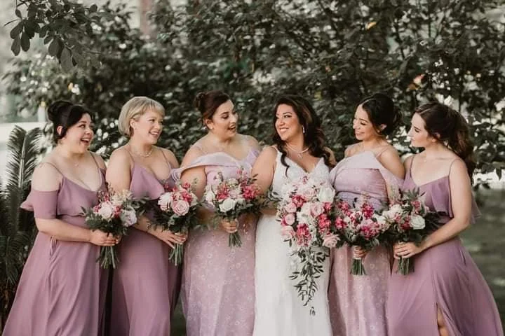 Six women in formal dresses standing outdoors, holding bouquets of flowers, and smiling at each other.