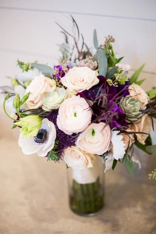 A bouquet of mixed flowers in a glass vase on a table, featuring white ranunculus, anemones, and purple and green accents.