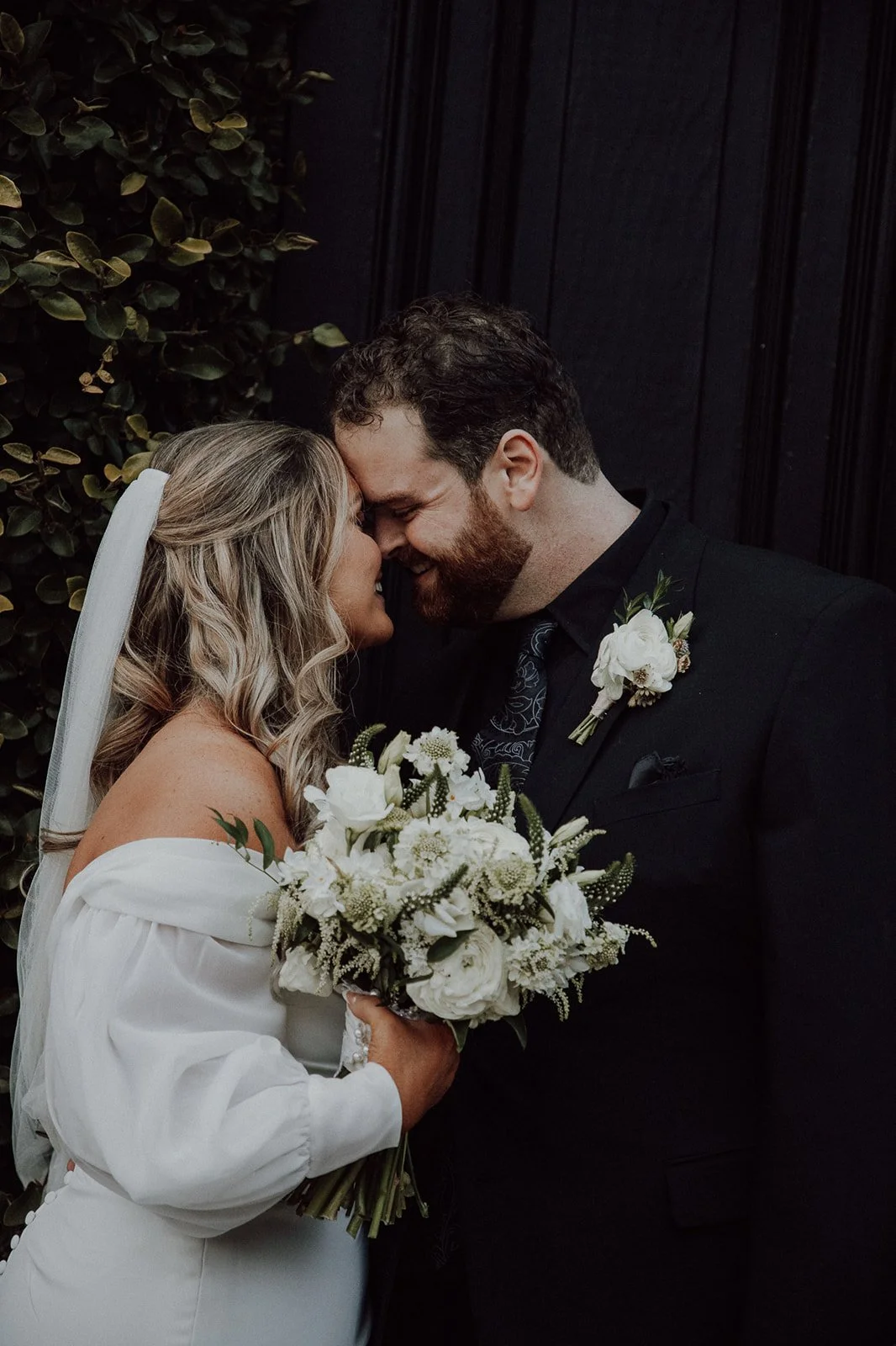 A bride and groom sharing a tender moment, touching their foreheads together, with the bride holding a bouquet of white flowers.