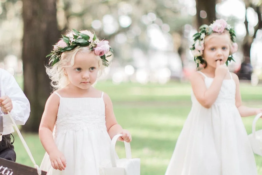 Two young girls wearing white dresses and floral crowns in a park.