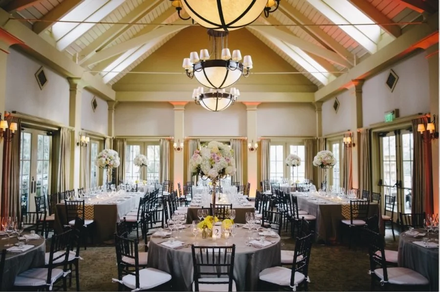 Elegant banquet hall with high vaulted ceiling, large chandeliers, and multiple round tables set for a formal event with floral centerpieces and place settings.
