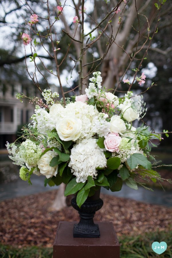 A bouquet of white and pale pink flowers, including roses and hydrangeas, placed in a black vase outdoors with trees and a house in the background.
