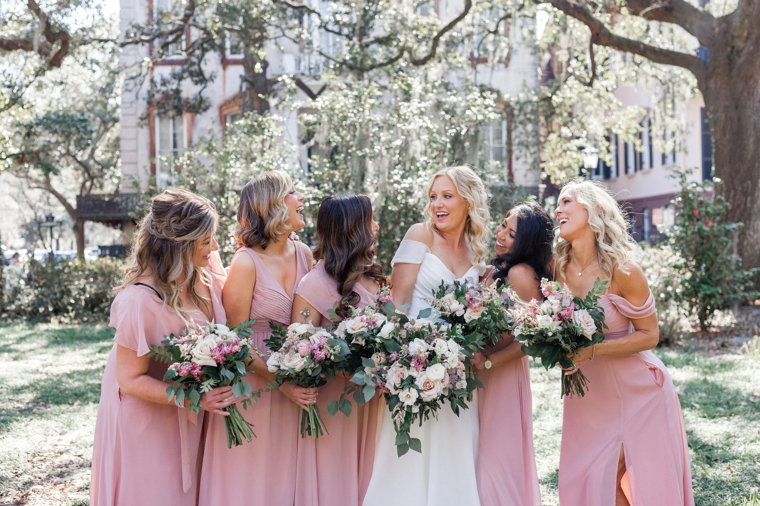 A group of six women, one in a white wedding dress and five in pink bridesmaid dresses, standing outdoors and sharing a joyful moment with bouquets of flowers in their hands.