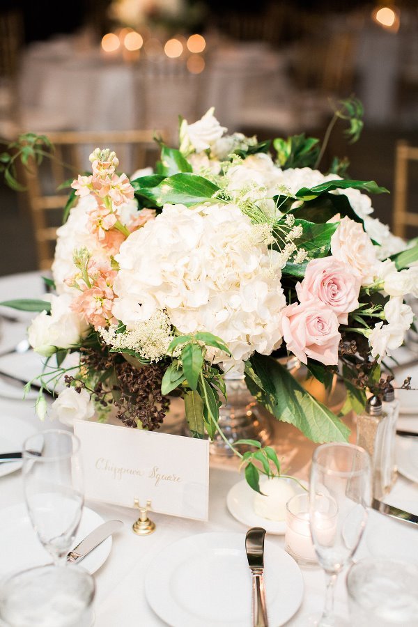 Elegant table centerpiece with white and blush pink roses and hydrangeas, surrounded by glassware and table setting for a formal event.