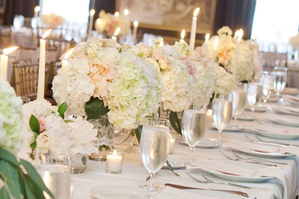 Elegant wedding reception table with white hydrangea and other floral centerpieces, tall lit candles, and glassware on white tablecloths.