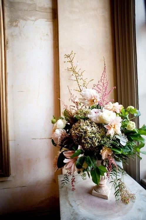 A large floral arrangement in a cream-colored vase, featuring white peonies, hydrangeas, and pink lilies, placed on a marble surface near a window with beige curtains.