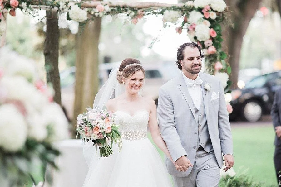 A bride and groom hold hands during their wedding ceremony outdoors under an arch decorated with flowers. The bride is smiling and holding a bouquet of pink and white flowers. The groom is dressed in a light gray suit with a white shirt and bow tie. 
