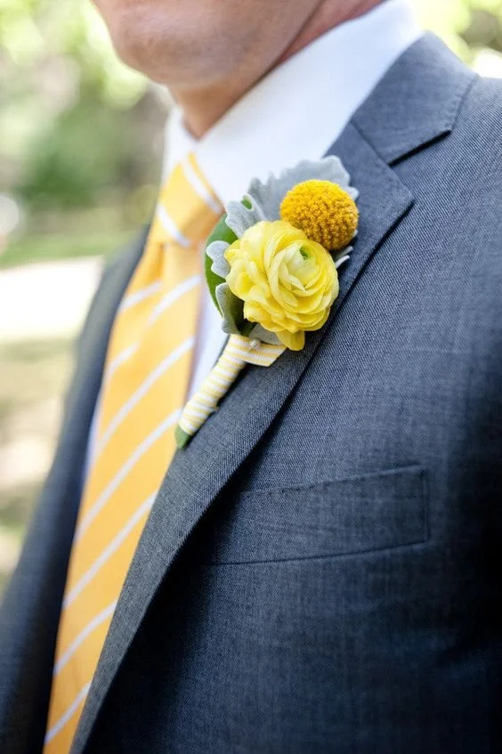 A man wearing a dark gray suit and a yellow striped tie with a boutonniere of yellow flowers and greenery on his lapel.