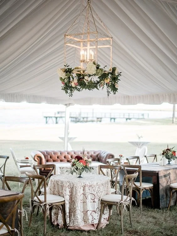 Elegant event setup under a tent with a chandelier, floral centerpieces, a pink tufted sofa, and round tables with lace tablecloths and chairs.