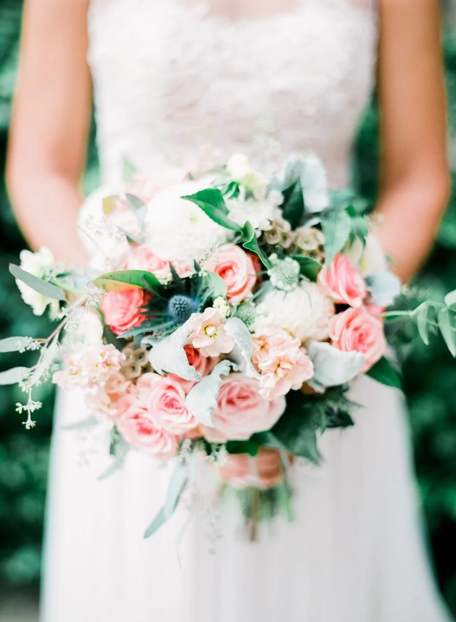 Bride holding a bouquet of pink roses, green leaves, and various flowers, in an outdoor setting.