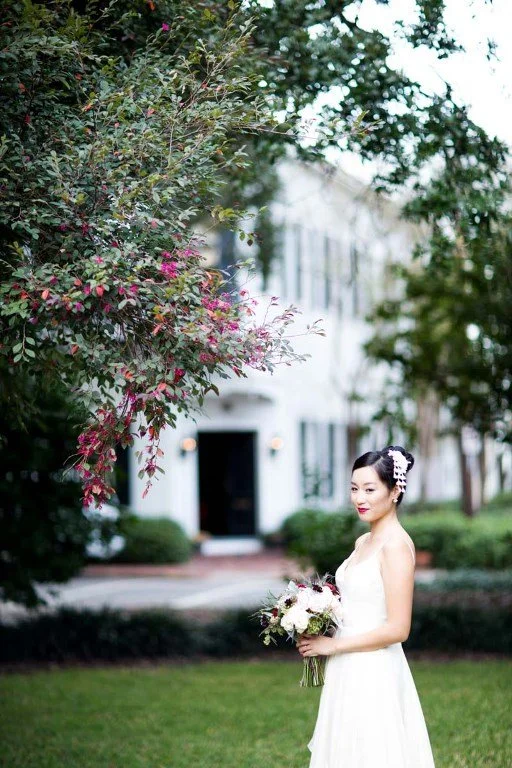 A bride in a white wedding dress holding a bouquet of flowers outdoors, standing on a grassy lawn with a large tree and a white building in the background.