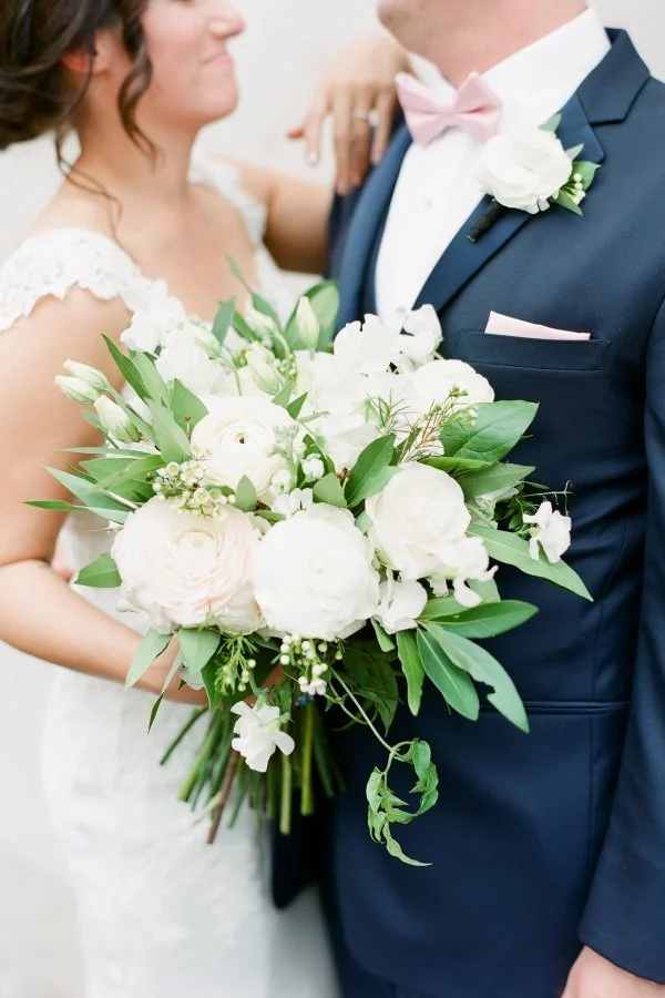 Bride and groom at their wedding, bride holding a large bouquet of white flowers, groom wearing a navy suit with pink bow tie and boutonniere.