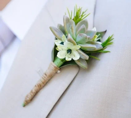 A small boutonniere with green and white succulent and flower, wrapped in twine, resting on a light-colored surface.