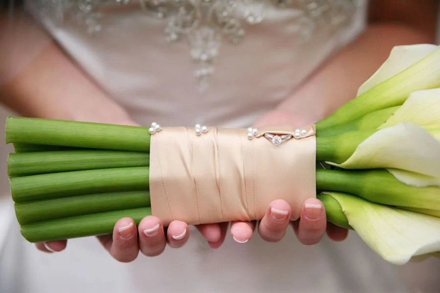 Person holding a bouquet of white calla lilies wrapped with beige satin ribbon and pearl pins, wearing a wedding dress.