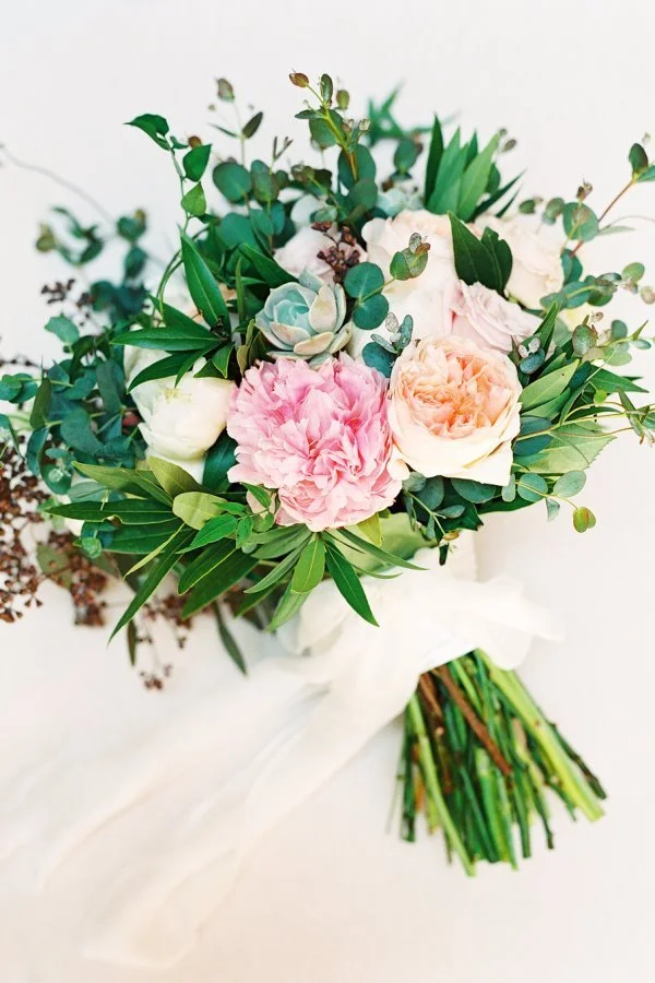 A bouquet of pink, white, and light peach roses and peonies with green leaves and eucalyptus, wrapped with a white ribbon.