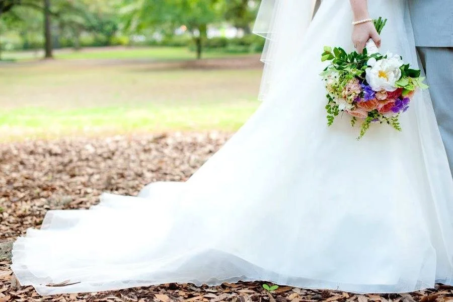 Close-up of a bride holding a colorful flower bouquet outdoors during a wedding, with part of her white wedding gown visible.